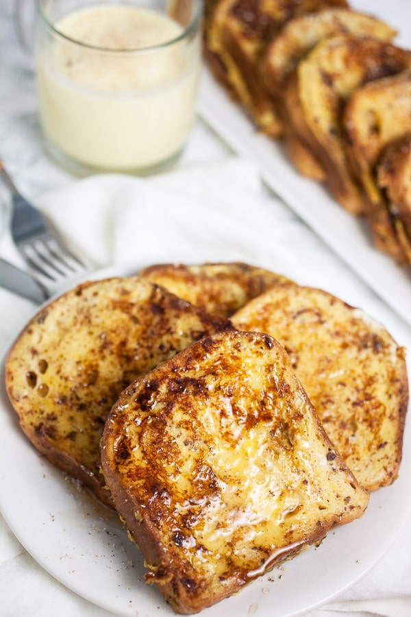 This Eggnog French Toast is a super easy breakfast recipe for Christmas morning and the holidays! Gluten free bread is soaked in a cinnamon egg mixture and then toasted until golden. It's topped with warm maple syrup and nutmeg. Everyone will love this French Toast! #breakfast #eggnog #christmas #glutenfree French toast on white plate with glass of eggnog.