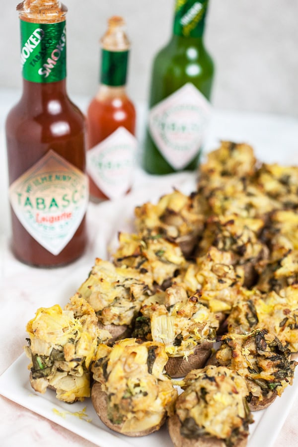 Spinach artichoke stuffed mushrooms on white serving platter in front of three varieties of Tabasco sauce.