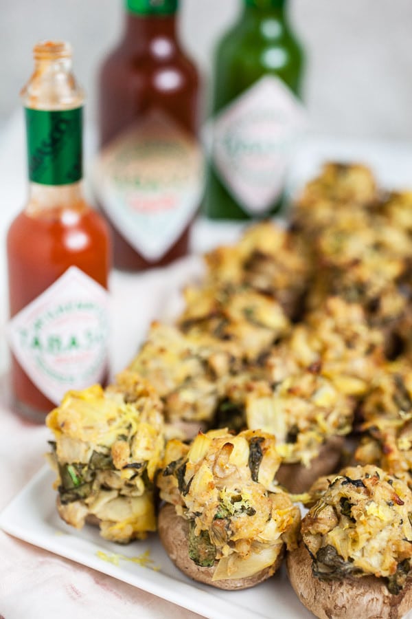 Spinach artichoke stuffed mushrooms on white serving platter in front of three varieties of Tabasco sauce.