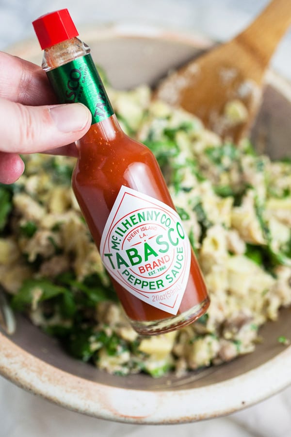 Fingers holding bottle of Tabasco sauce in front of spinach artichoke filling in ceramic bowl.