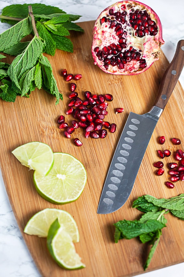 Pomegranate fruit and arils, sliced limes, and fresh mint on wooden cutting board with knife.
