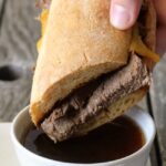 Slow Cooker Beef Sandwich Being Dipped Into Au Jus Sauce In Small White Bowl