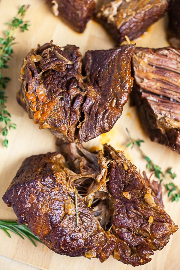 Cooked beef chuck roast on wooden cutting board with rosemary and thyme.