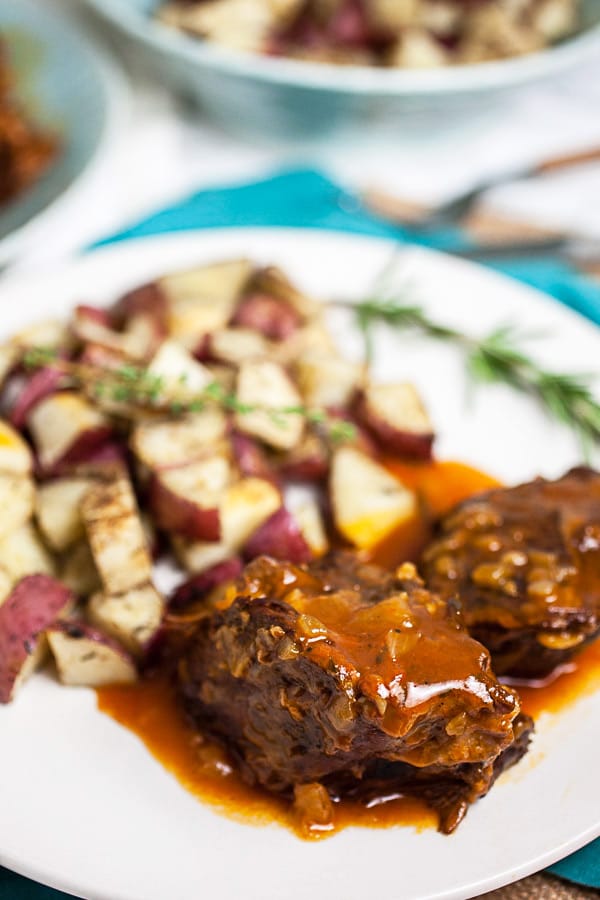 Beef pot roast with gravy, roasted potatoes, and rosemary on white plate.