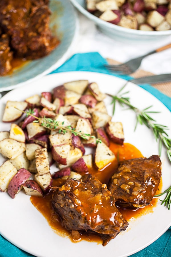 Pot roast with gravy, roasted potatoes, and rosemary on white plate.