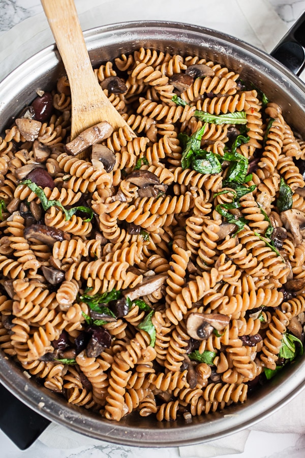 Mushroom olive rotini pasta with fresh basil in skillet with wooden spoon.