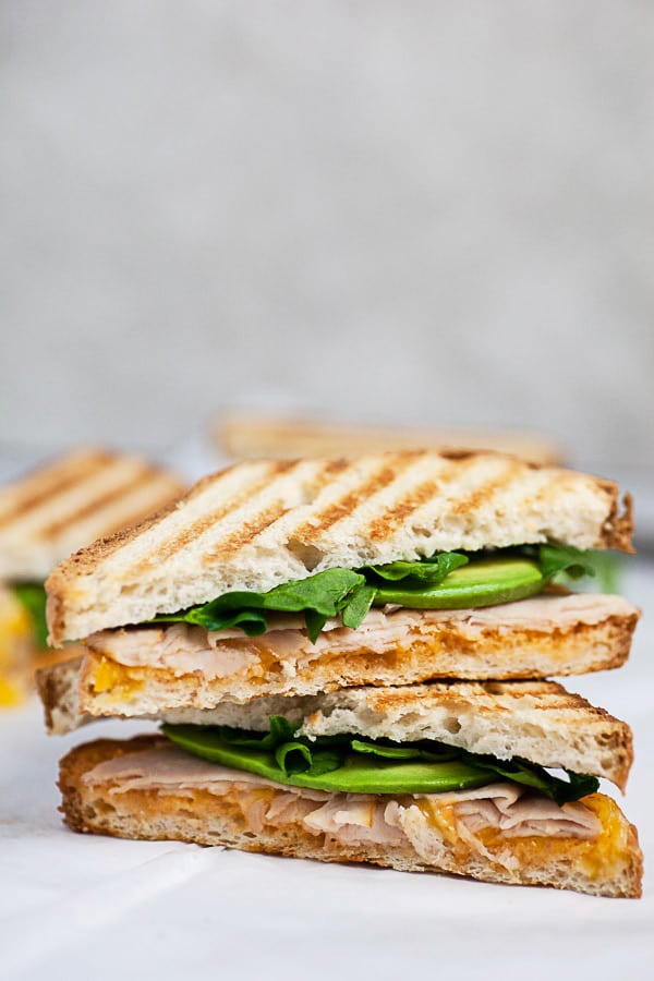 Chicken panini sandwich with avocado and spinach cut in half and stacked on white surface.