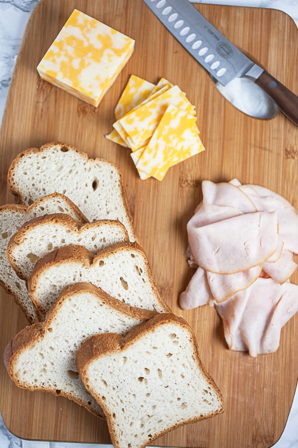 Sliced bread, cheese, and deli chicken on wooden cutting board with knife.