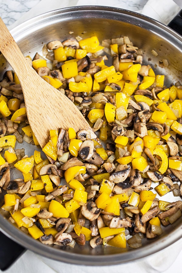 Diced mushrooms and yellow bell peppers sautéed in skillet with wooden spatula. 