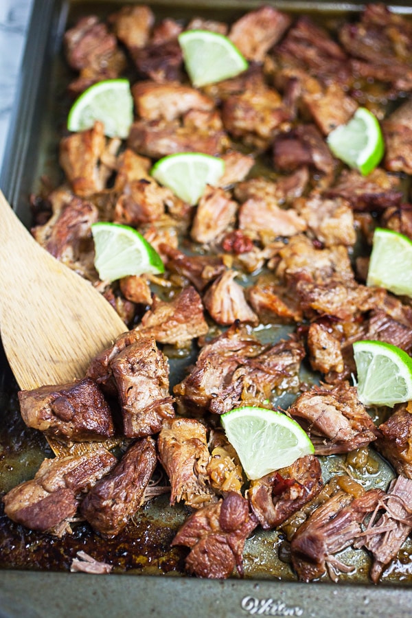Cooked shredded pork shoulder broiled on metal sheet pan with chunks of fresh lime.
