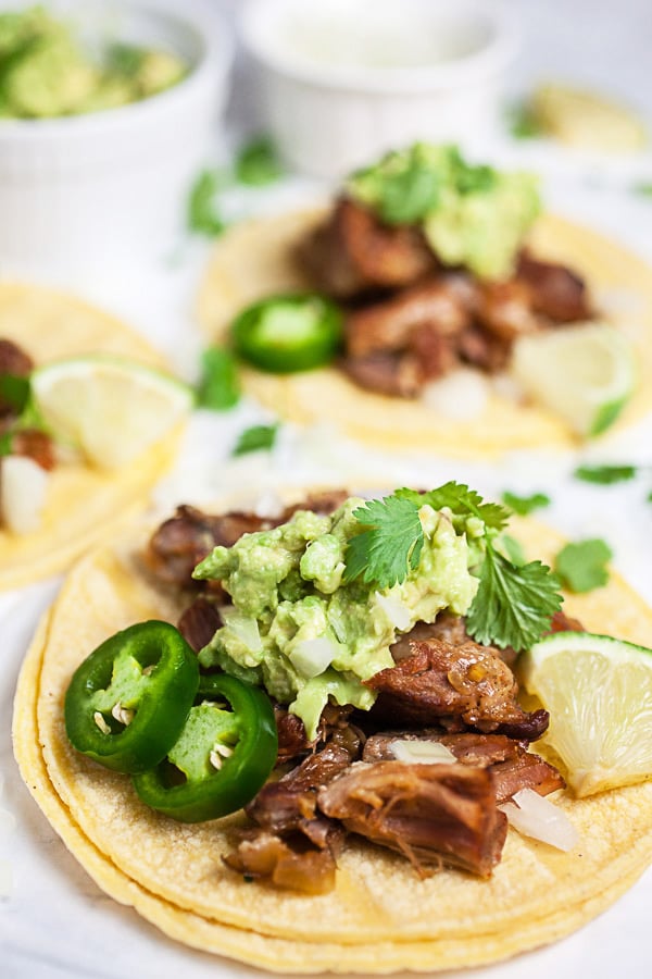 Instant Pot pork carnitas tacos on corn tortillas with guacamole, cilantro, jalapeno peppers, and limes.
