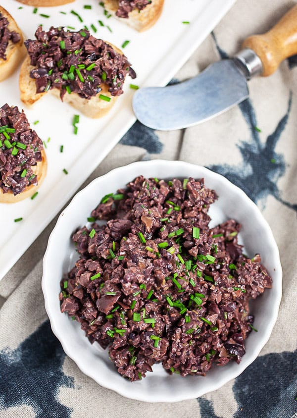 Small white bowl of olive tapenade next to serving tray of crostini.
