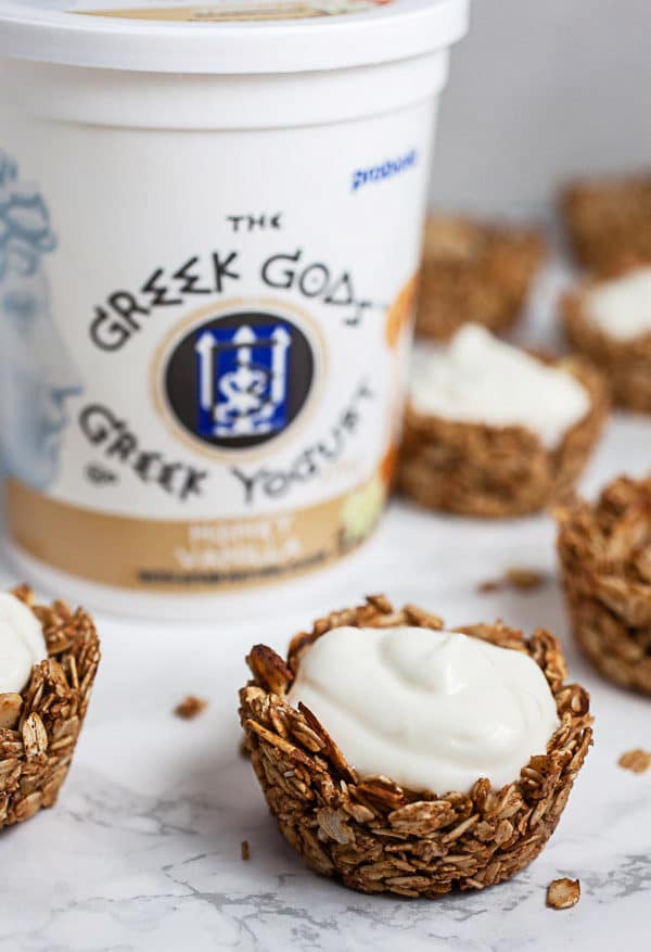 #ad These Greek Yogurt Oatmeal Cups are the perfect easy and healthy breakfast recipe! The oats are mixed with coconut oil, lemon zest, and cinnamon and then baked until crispy. They're filled with yogurt that's creamy and lightly sweetened with honey. Great for a brunch, dessert, or to serve at a party! @GreekGodsYogurt #oatmeal #yogurt #cups #healthy #glutenfree Baked oatmeal cups with yogurt and tub of Greek Gods yogurt on white surface.