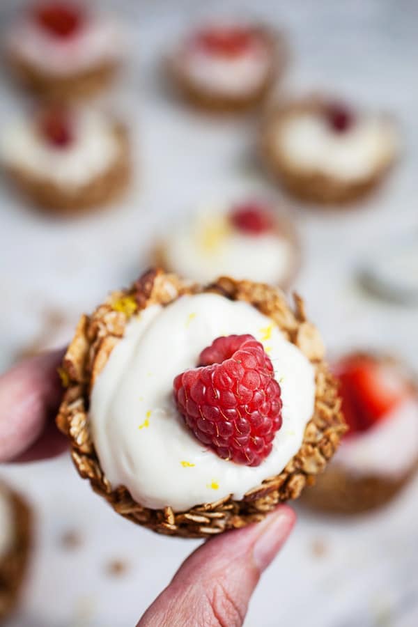 #ad These Greek Yogurt Oatmeal Cups are the perfect easy and healthy breakfast recipe! The oats are mixed with coconut oil, lemon zest, and cinnamon and then baked until crispy. They're filled with yogurt that's creamy and lightly sweetened with honey. Great for a brunch, dessert, or to serve at a party! @GreekGodsYogurt #oatmeal #yogurt #cups #healthy #glutenfree Fingers holding oatmeal yogurt cup with fresh raspberry.