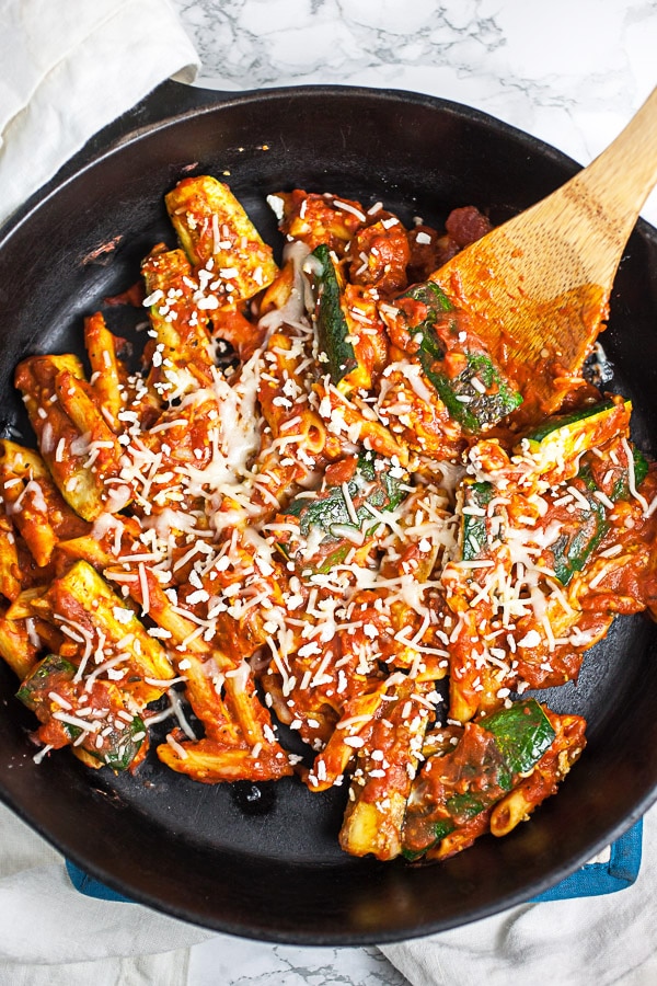 This Skillet Zucchini Parmesan recipe makes the perfect easy and healthy weeknight dinner for one! The pasta is cooked in a rich tomato sauce, combined with fresh zucchini, and then baked in the oven. It's topped with mozzarella and Parmesan cheese. This meal is vegetarian and gluten free! #pasta #zucchini #skillet #vegetarian #glutenfree Zucchini Parmesan in cast iron skillet with wooden spoon.