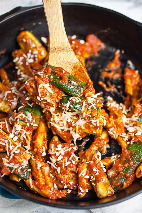 This Skillet Zucchini Parmesan recipe makes the perfect easy and healthy weeknight dinner for one! The pasta is cooked in a rich tomato sauce, combined with fresh zucchini, and then baked in the oven. It's topped with mozzarella and Parmesan cheese. This meal is vegetarian and gluten free! #pasta #zucchini #skillet #vegetarian #glutenfree Scoop of zucchini Parmesan lifted from cast iron skillet with wooden spatula.