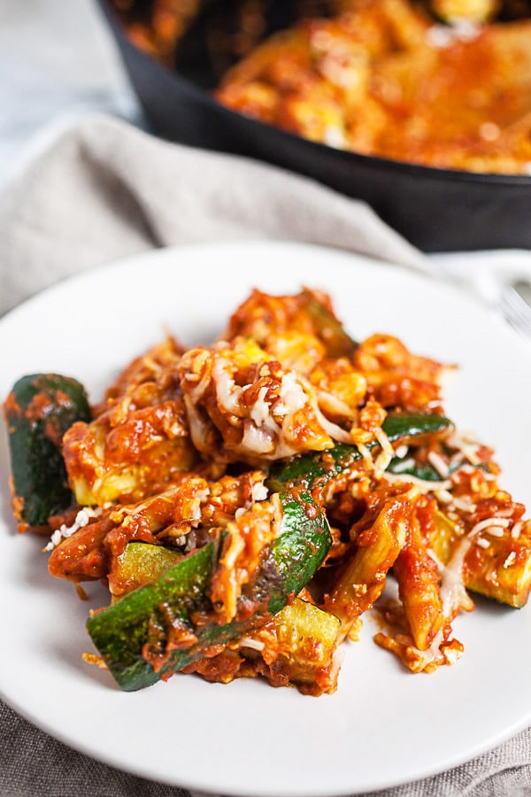 This Skillet Zucchini Parmesan recipe makes the perfect easy and healthy weeknight dinner for one! The pasta is cooked in a rich tomato sauce, combined with fresh zucchini, and then baked in the oven. It's topped with mozzarella and Parmesan cheese. This meal is vegetarian and gluten free! #pasta #zucchini #skillet #vegetarian #glutenfree Skillet zucchini Parmesan on small white plate in front of cast iron skillet.