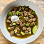 Chili verde in white bowl garnished with sour cream, diced avocadoes, and lime wedge.