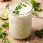 Cilantro ranch dressing in small glass Mason jar next to lime wedges.