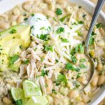 White chicken chili in white bowl with spoon garnished with sour cream, avocado, lime, and cilantro.