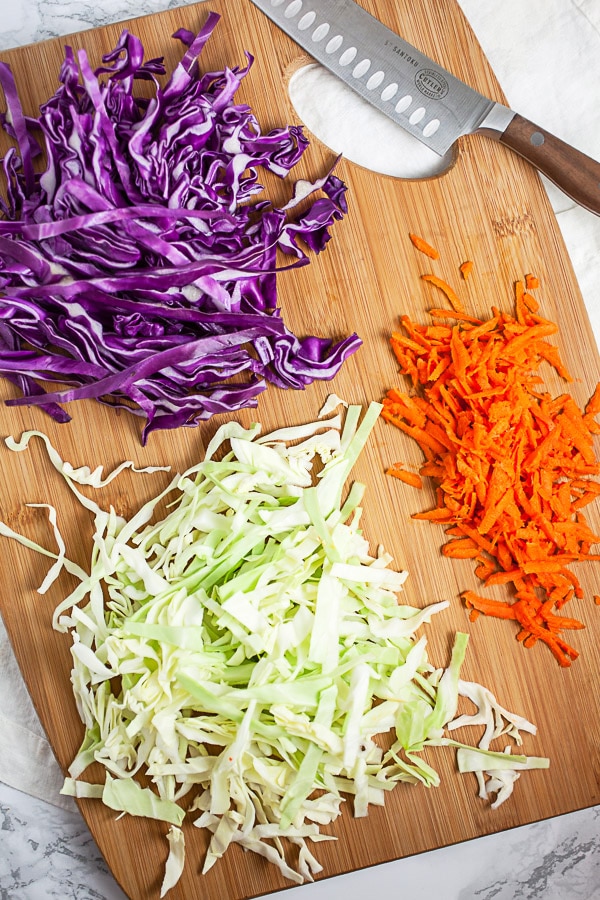 Shredded red and green cabbage and carrots on wooden cutting board with knife.