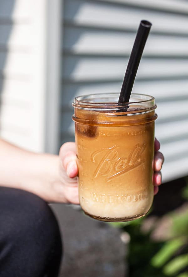 Hand holding glass mason jar of coffee with almond milk outdoors.