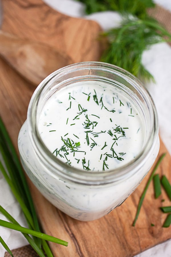 Homemade ranch dressing with fresh herbs in glass jar on wooden serving board.