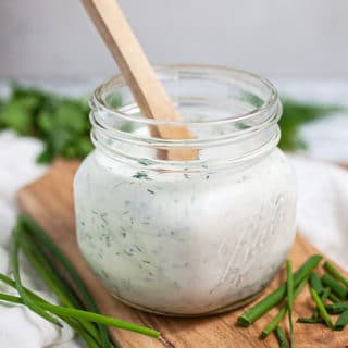 Homemade ranch dressing in glass jar with fresh herbs on wooden serving board.