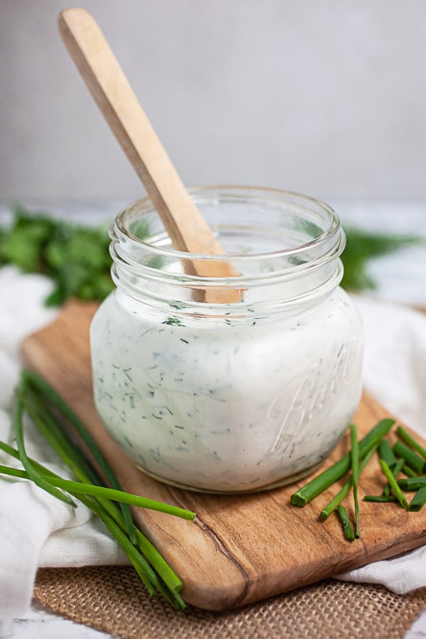 Homemade ranch dressing in glass jar with fresh herbs on wooden serving board.