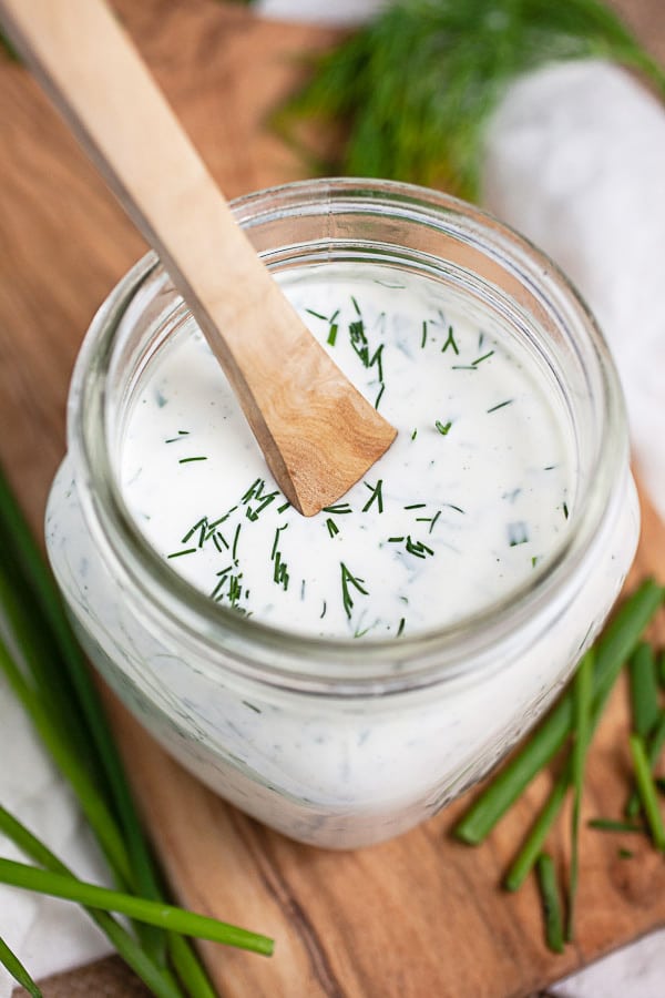 Wooden spoon in glass jar of ranch dressing on wooden serving board with fresh herbs.