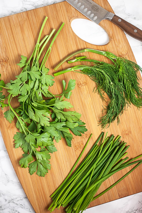 Fresh parsley, dill, and chives on wooden cutting board with knife.