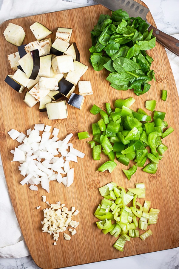 Minced garlic, onions, banana peppers, green bell peppers, eggplant, and spinach on wooden cutting board with knife.