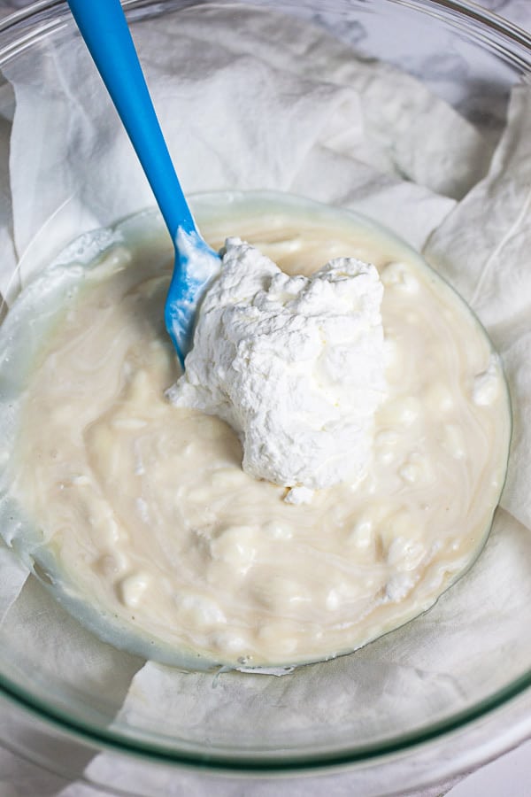 Scoop of whipped cream added to sweetened condensed milk in glass bowl with blue spatula.