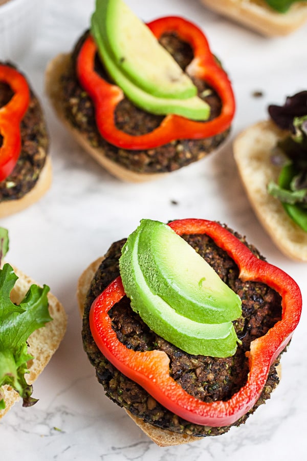 Open faced black bean mushroom burgers on buns with red bell pepper rounds and sliced avocado.