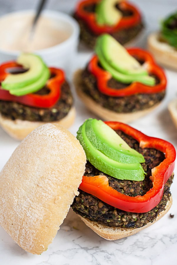 Black bean mushroom burgers on buns with red bell pepper rounds, sliced avocado, and bowl of chipotle mayo.