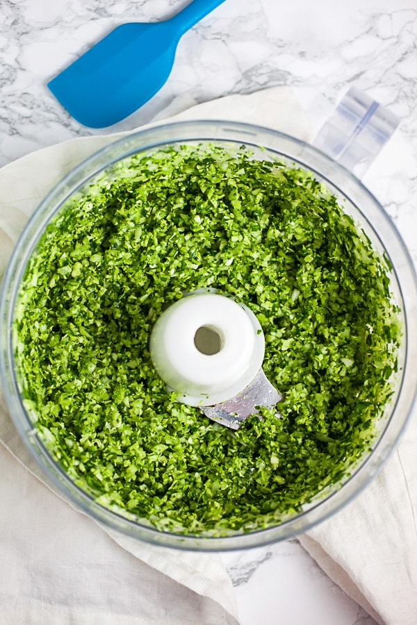 Broccoli and onions minced together in food processor next to blue spatula.