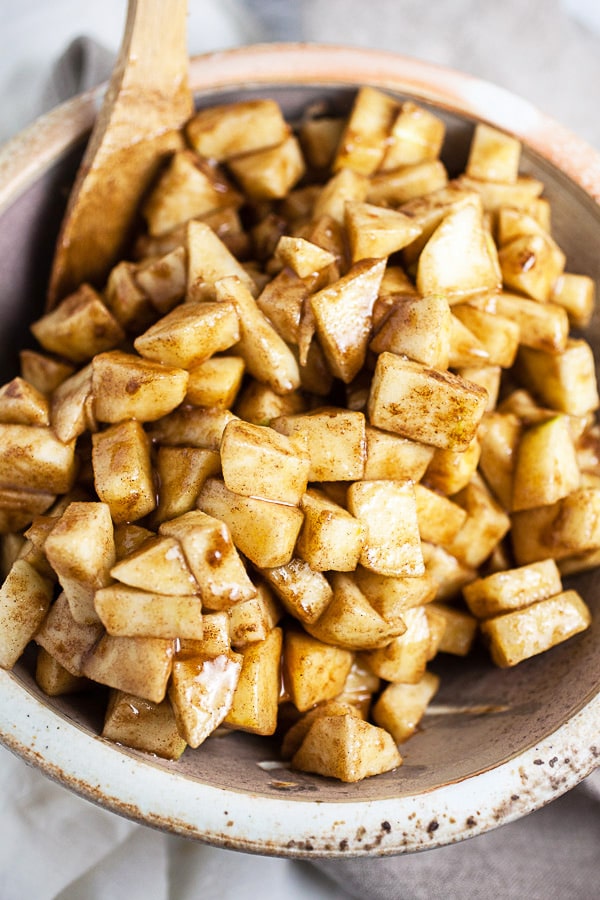 Diced apples with brown sugar and spices in ceramic bowl with wooden spoon.