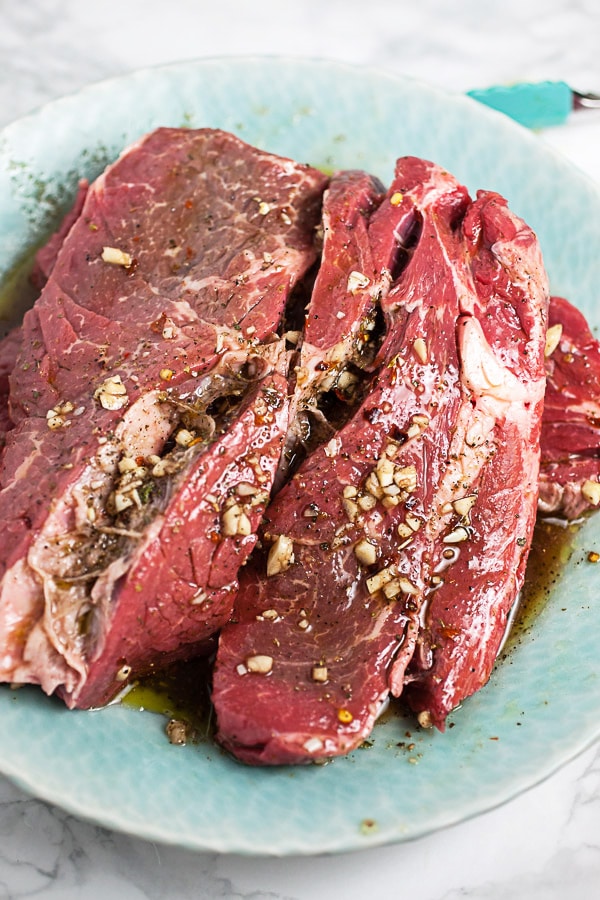 Chuck steaks marinating in olive oil, vinegar, garlic, and spices in blue bowl.