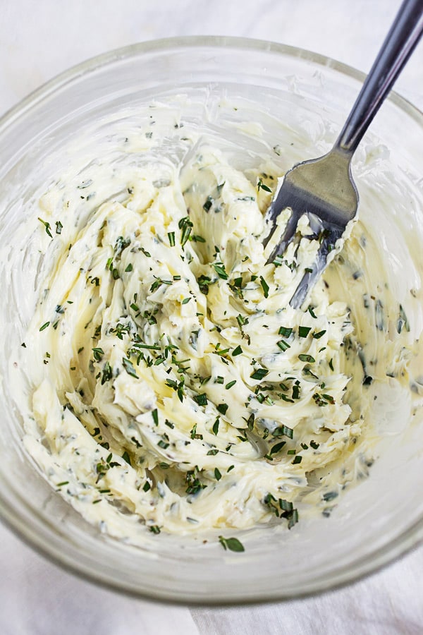 Compound butter with garlic, thyme, and rosemary in small glass bowl with fork.