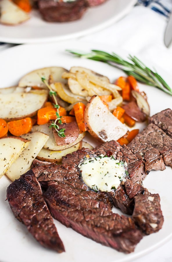Chuck steak with compound butter on white plate with potatoes, carrots, and rosemary.