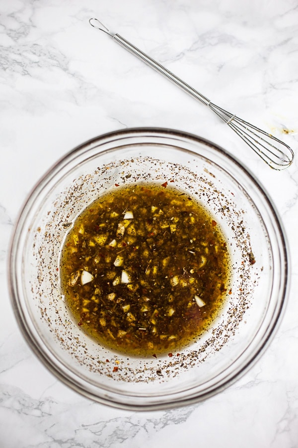 Olive oil, vinegar, garlic, and Italian spice marinade in small glass bowl next to whisk.