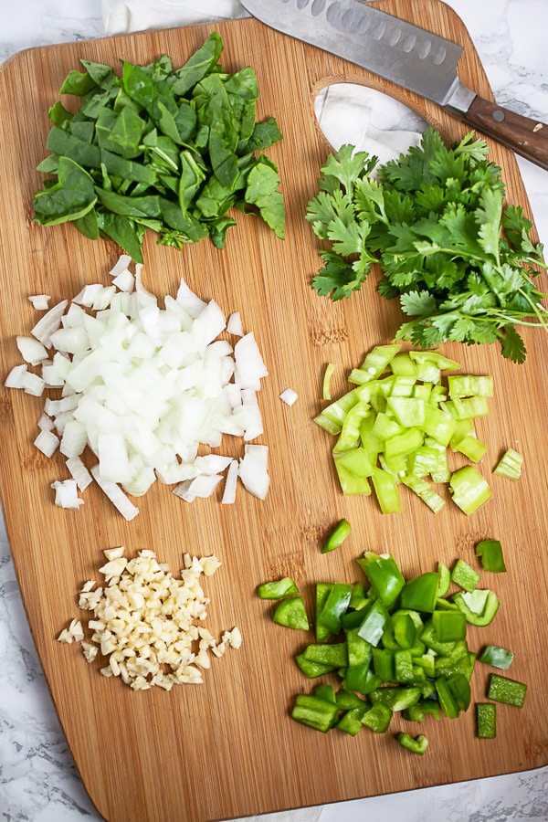 Minced garlic, onions, green bell peppers, banana peppers, spinach, and cilantro on wooden cutting board with knife.