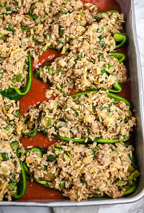 Uncooked halved poblano peppers with filling and sauce in metal cake pan.