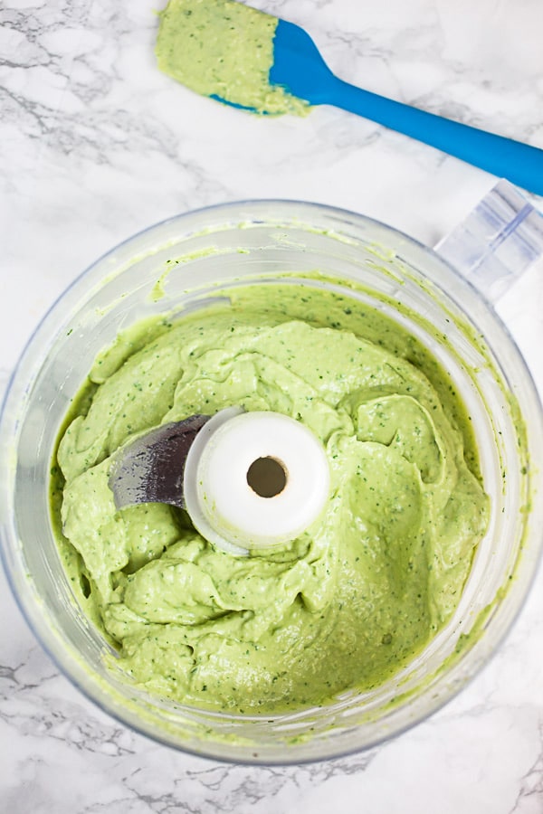 Avocado cream sauce in food processor next to blue spatula.