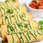 Baked chicken taquitos drizzled with avocado cream sauce on white serving platter.