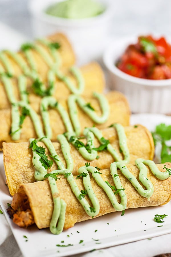 Baked chicken taquitos drizzled with avocado cream sauce on white serving platter.