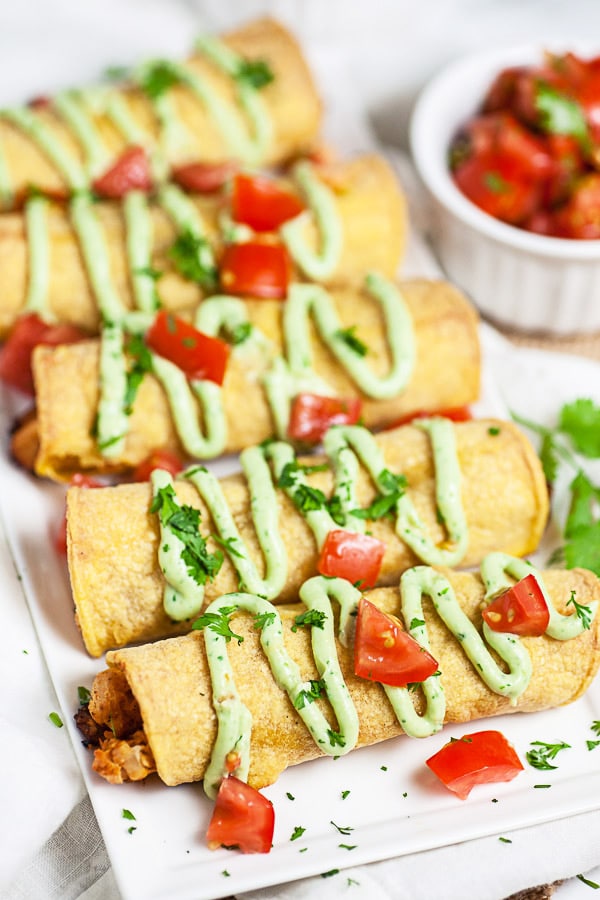 Baked chicken taquitos on white serving platter garnished with avocado cream sauce and diced tomatoes.