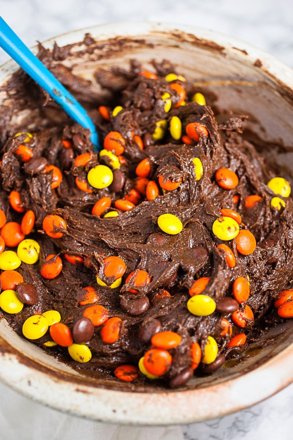 Chocolate cookie batter mixed with Reese's Pieces candies in ceramic bowl.