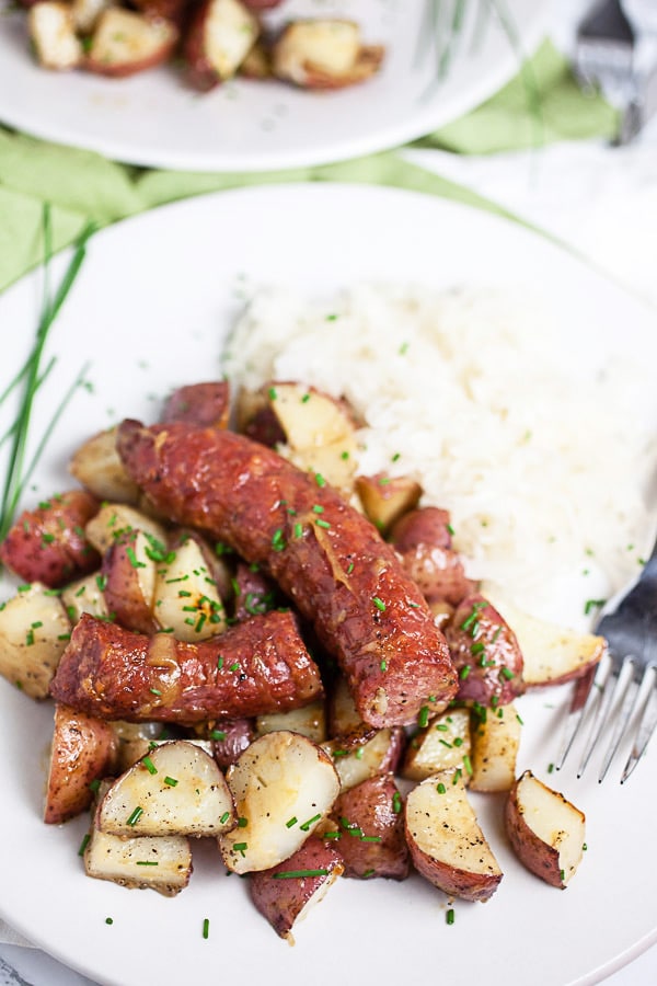 Sausage and potatoes garnished with fresh chives with sauerkraut on white plate.