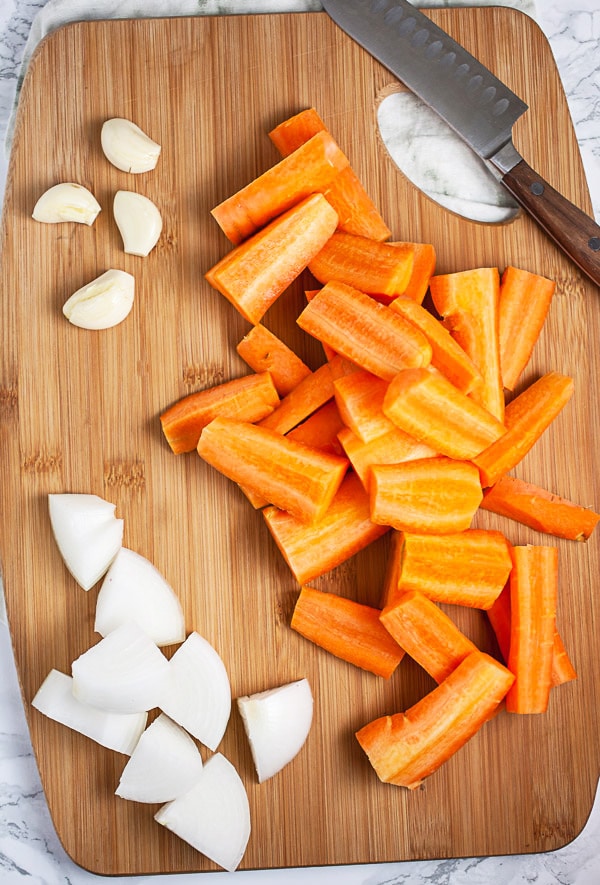 Chopped carrots, onion chunks, and garlic cloves on wooden cutting board with knife.